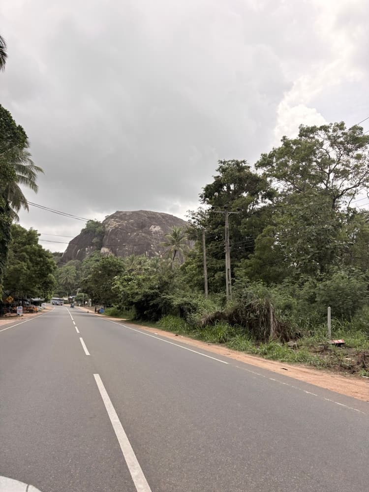 Quiet two-lane A6 road near Inamaluwa with a granite rock outcrop and trees beside it
